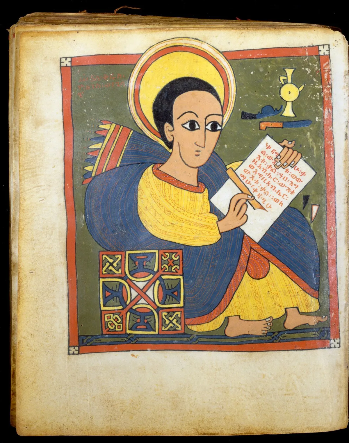 A manuscript painting of a haloed man in a blue and yellow robe writing on a scroll, with a decorated panel beneath.